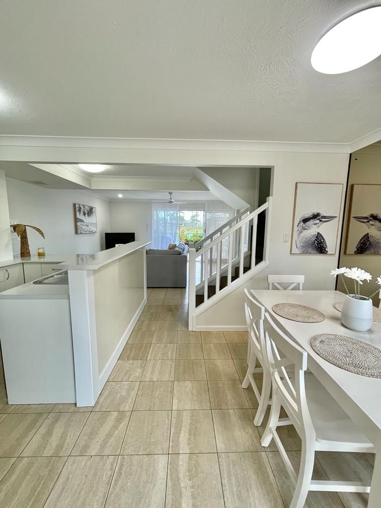 Une cuisine moderne avec des meubles blancs, un coin repas avec des chaises blanches et un escalier menant à un balcon.