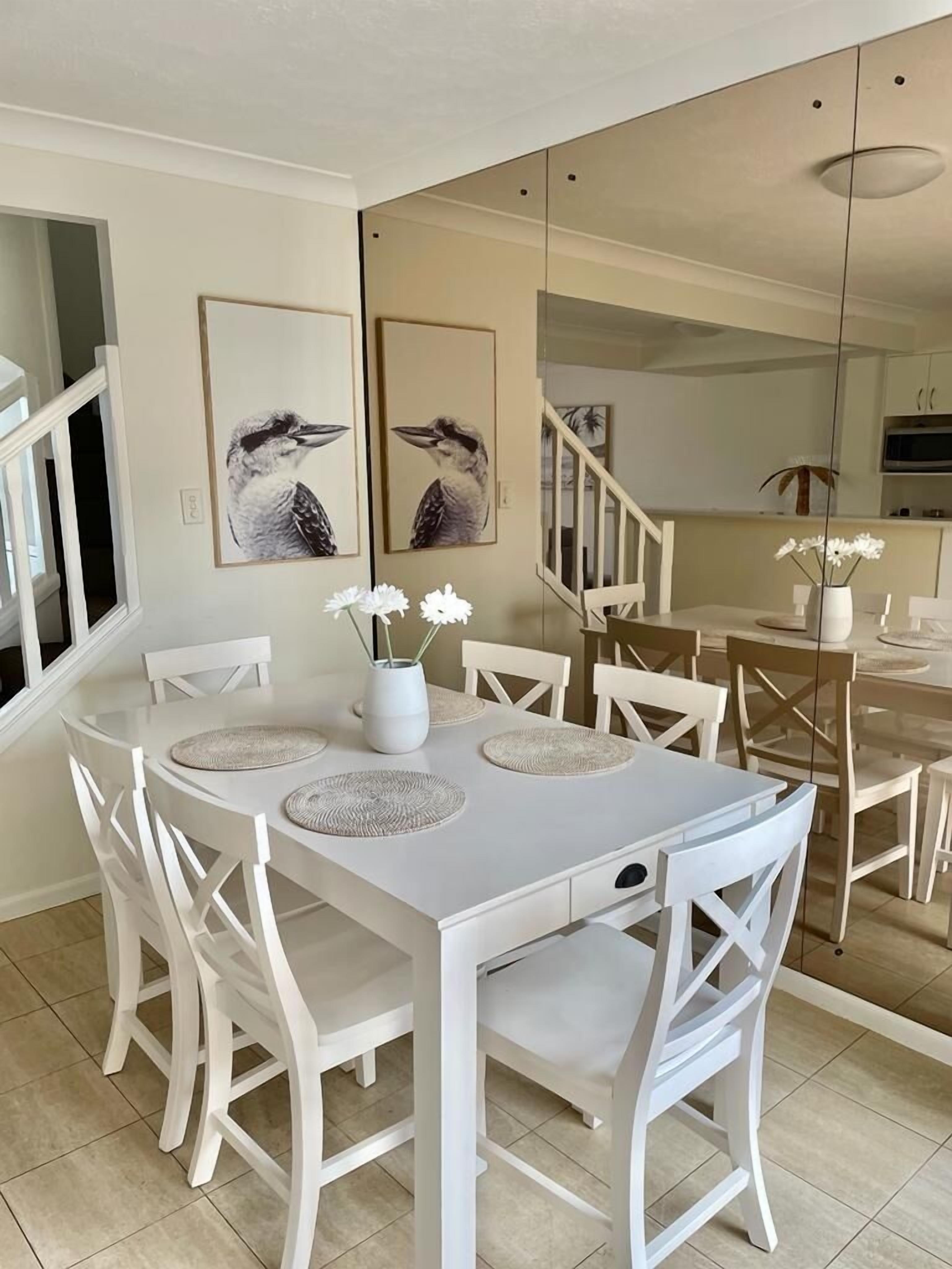 A dining area with a white table and chairs, two framed bird paintings on the wall, and a vase with flowers on the table.
