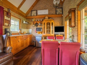 A rustic kitchen with wooden cabinets, a dining table with red chairs, a refrigerator, and a microwave.