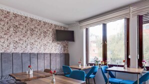 A modern restaurant with wooden tables and blue chairs, a grey cushioned bench, and a floral wallpaper wall. - Cozy Corner in Oberhausen (Oberhausen)