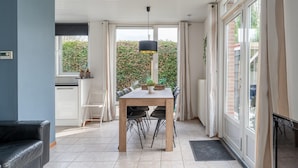 A modern dining area with a wooden table and chairs, a hanging pendant light, and a view of a garden through large windows. - Holiday Home in Zeeland Near Lake (Baarland)