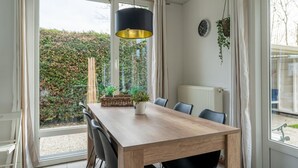A dining area with a wooden table, black chairs, a hanging pendant light, and a view of a garden through large windows. - Holiday Home in Zeeland Near Lake (Baarland)