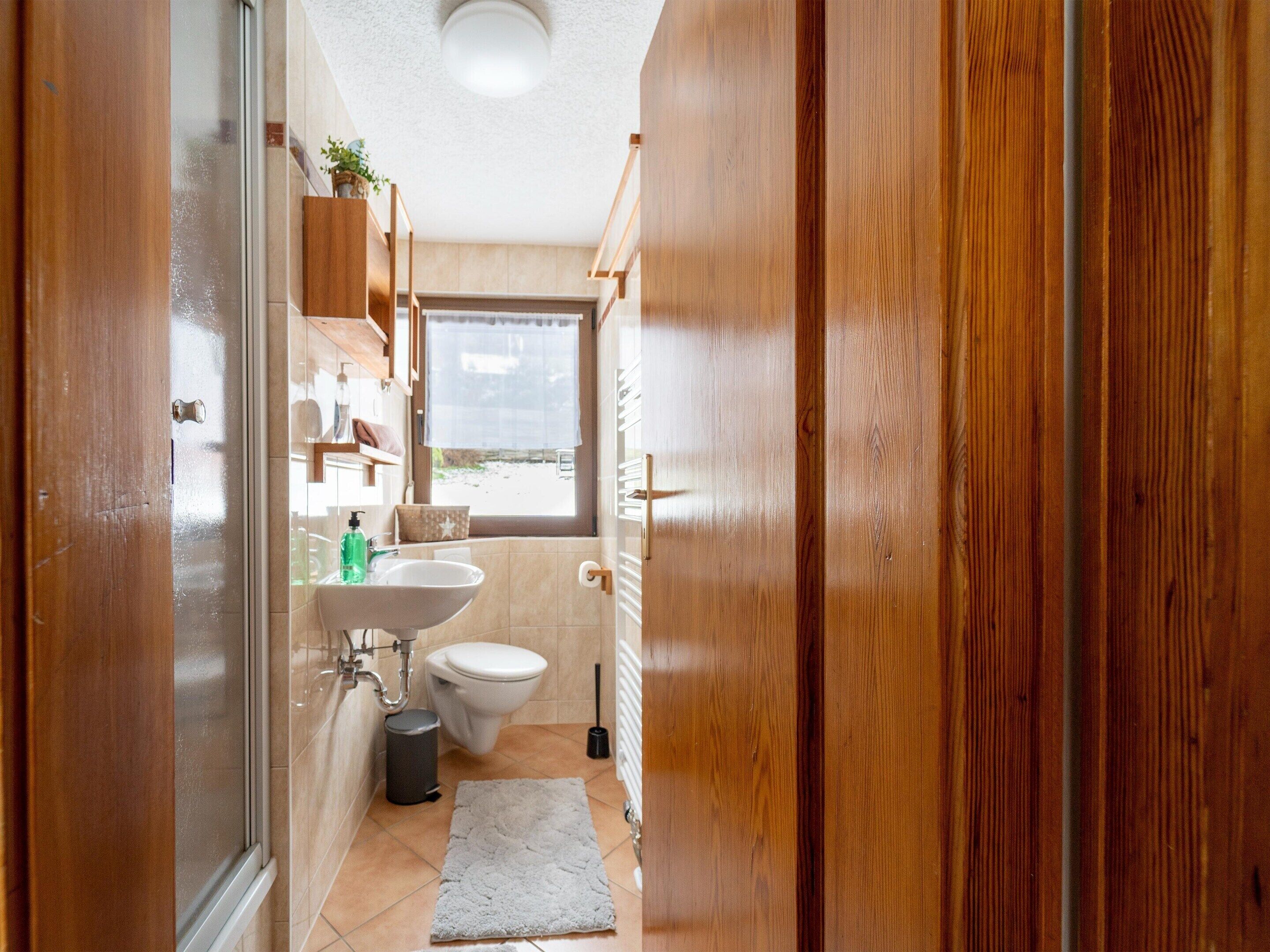 Une petite salle de bain avec une porte en bois, un lavabo blanc, des toilettes et une fenêtre donnant sur l’extérieur.