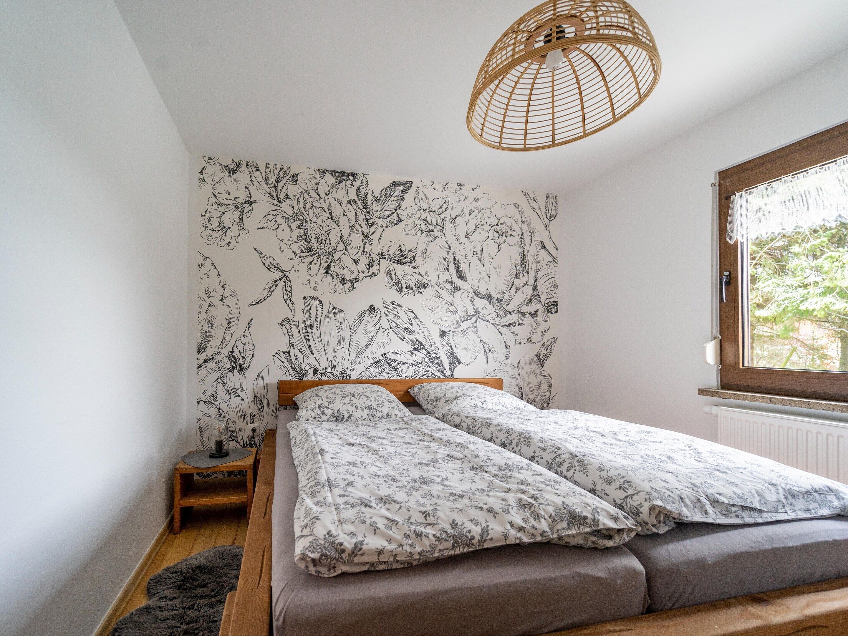 A bedroom with a bed, a nightstand, a small table, a wicker lamp, and a window with a view of greenery.