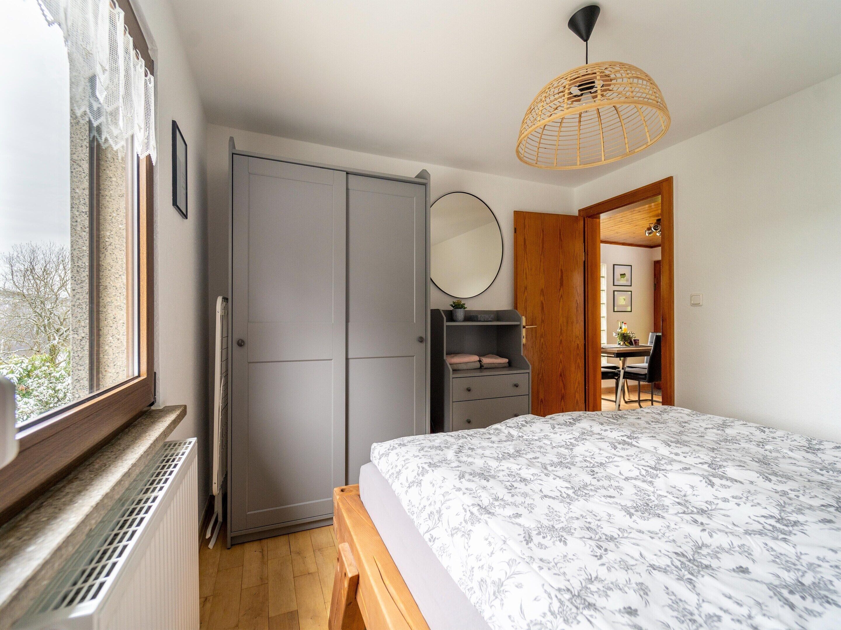 A bedroom with a large bed, a wardrobe, a mirror, and a window with a view of trees.