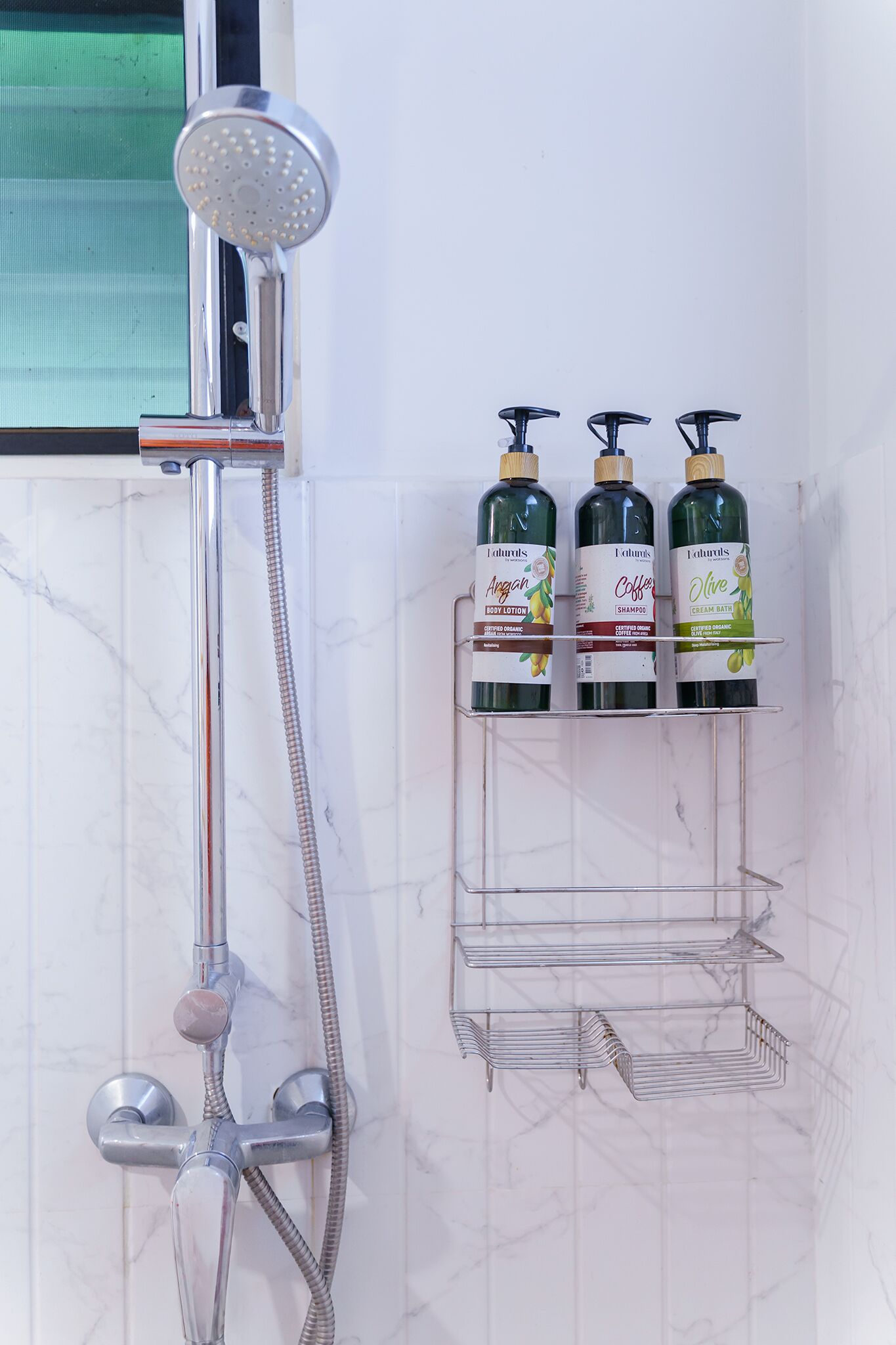 A bathroom with a shower, a hand dryer, and three bottles of body lotion on a shelf.