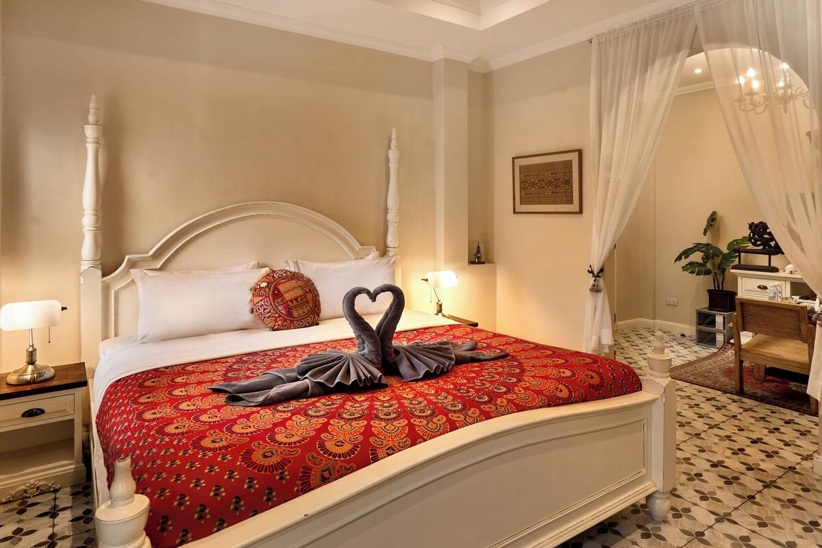 A neatly made bed with a red patterned bedspread and two swan-shaped towels.