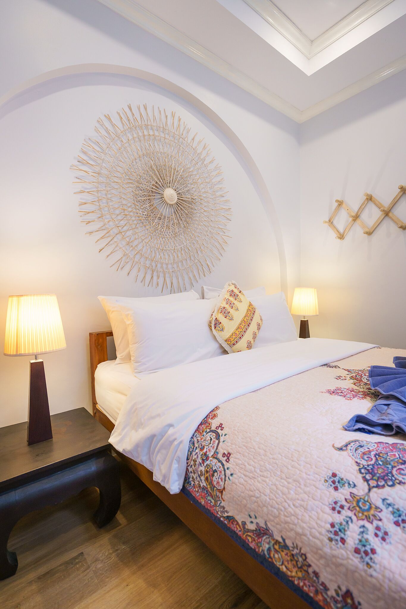 A neatly made bed with a patterned bedspread, two bedside lamps, a decorative wall piece, and a wooden bench.