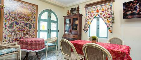 A dining area with a round table covered in a red tablecloth, surrounded by wicker chairs. There is a cabinet with glass doors and a colorful tapestry on the wall.