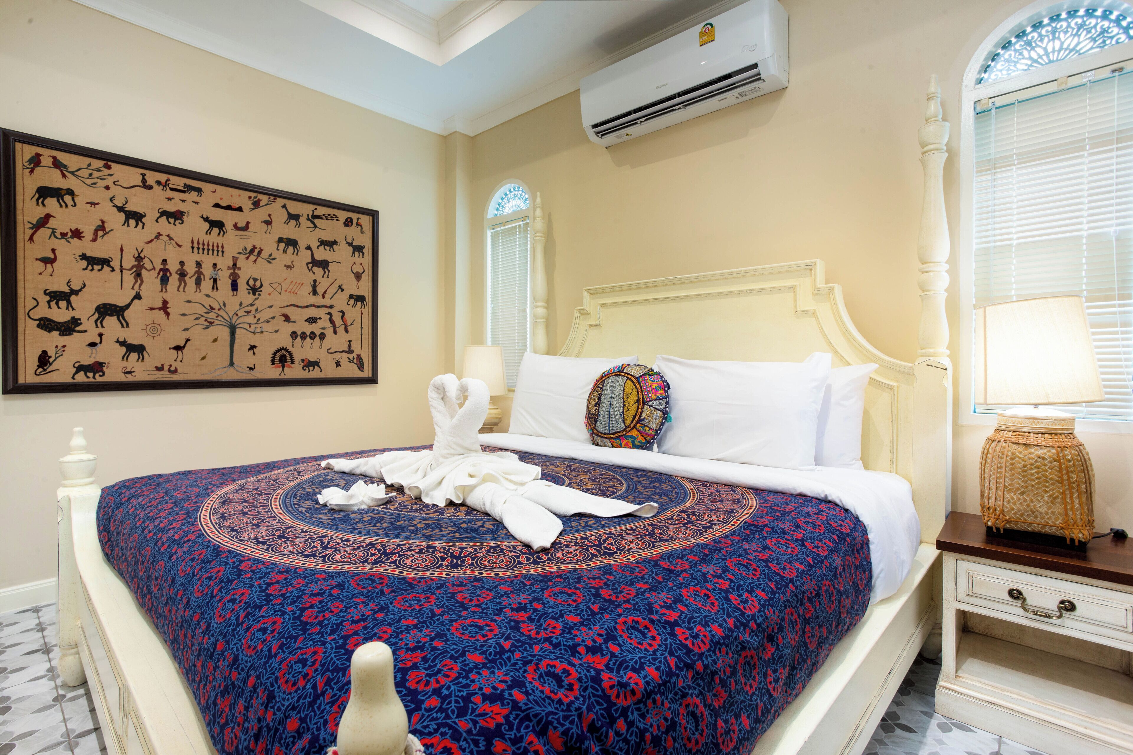 A hotel room with a large wall tapestry, a bed with a patterned bedspread, a decorative pillow, a nightstand with a lamp, and an air conditioning unit.