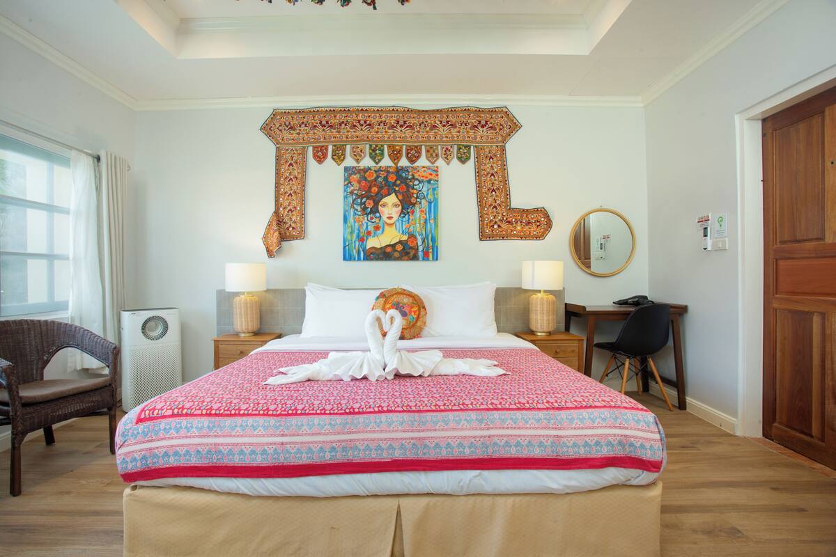 A hotel room with a large bed, two bedside tables with lamps, a chair, a desk, a wooden door, and a wall tapestry.