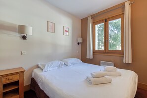 Uma cama bem arrumada com roupa de cama e toalhas brancas, uma mesa de cabeceira, uma luminária na parede e uma janela com cortinas.