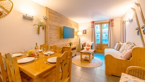 A cozy living room with a wooden dining table set for four, a sofa, and a TV mounted on the wall. - V Loriaz 14: Beside Ski Lift; Pool, Sauna & Gym, Vallorcine (Argentiere), France (Vallorcine (Argentiere))