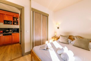 Habitación de hotel con cama de madera, una mesita de noche con lámpara y vista a la cocina a través de una puerta abierta.