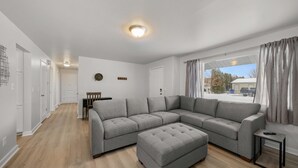 Een moderne woonkamer met een grijze modulaire bank, een passende voetenbankje, en een kleine bijzettafel. In de achtergrond is een eethoek met een tafel en stoelen zichtbaar.