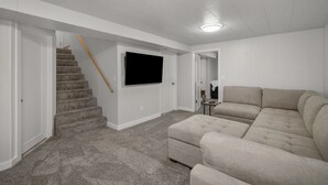 Een woonkamer met een beige modulaire bank, een flatscreen televisie aan de muur en een betegeld trappenhuis.