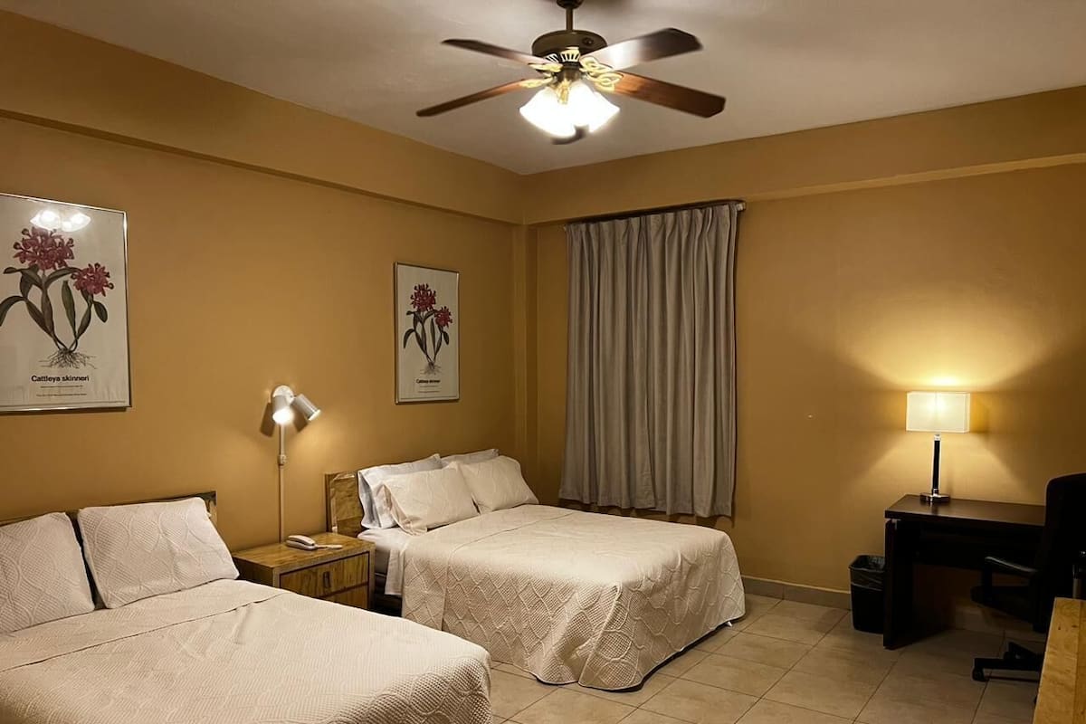 A hotel room with two beds, a desk, a chair, a ceiling fan, and two lamps.