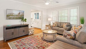 A living room with a sofa, coffee table, and a patterned rug. There is a TV mounted on the wall, a ceiling fan, and a window with blinds. - House is close to Beach Pier, Shops & Restaurants (Saint Simons Island)