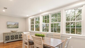A dining area with a wooden table and chairs, large windows with white frames, and a gray cabinet with a bowl on top. - House is close to Beach Pier, Shops & Restaurants (Saint Simons Island)