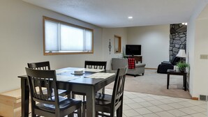 A dining area with a table and chairs, a living room with a sofa and a TV, and a stone accent wall. - Aksarben Two Bed, One Bath Duplex Suite (Omaha)