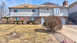 A two-story house with a front garage, a brick chimney, and a small front yard with shrubs and a tree. - Aksarben Two Bed, One Bath Duplex Suite (Omaha)