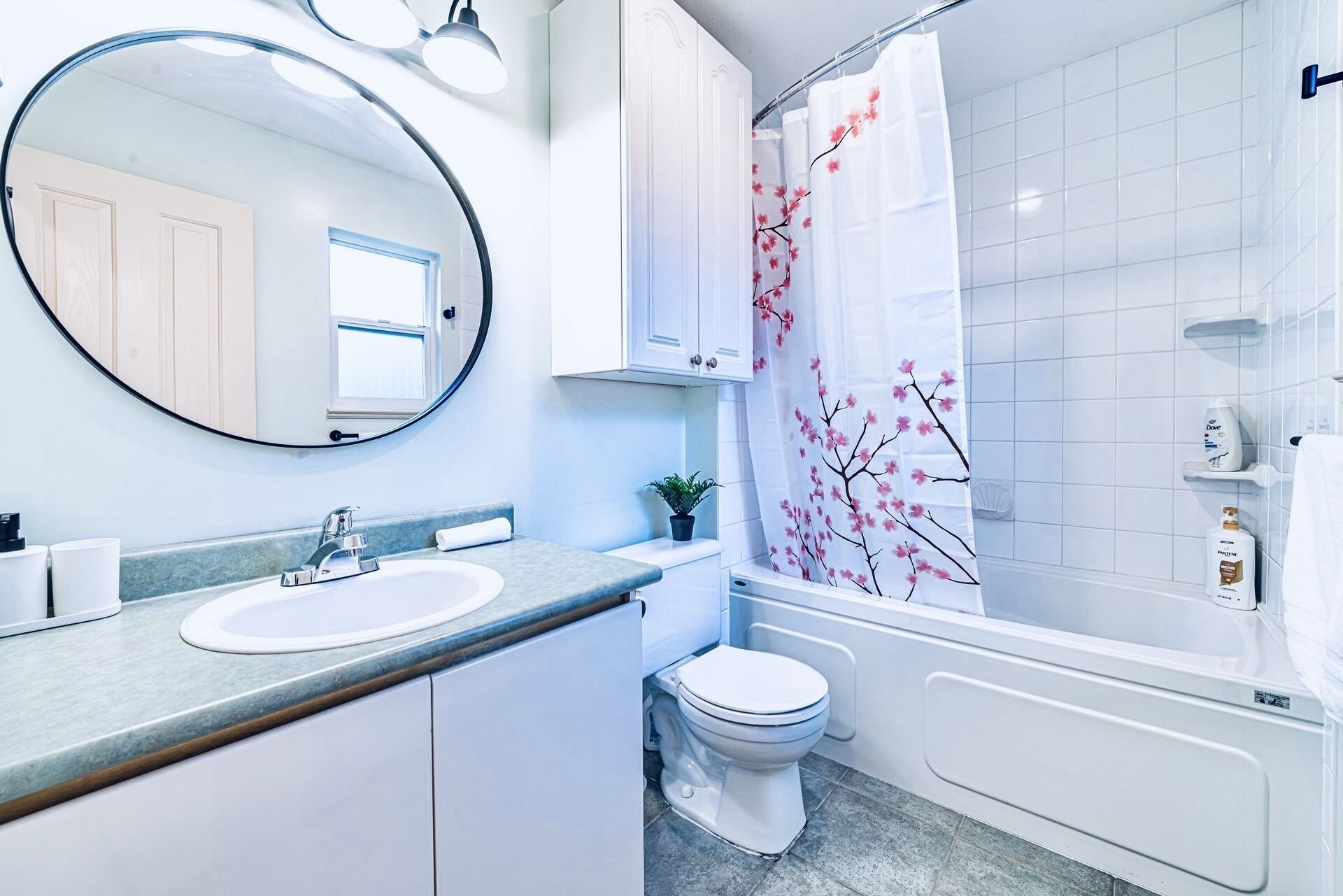 A modern bathroom with a round mirror, white cabinets, a sink, a toilet, and a shower with a floral shower curtain.