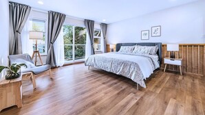 Ein Schlafzimmer mit einem großen Bett, einem Sessel, einem Nachttisch, einer Lampe und einem Fenster mit Vorhängen.