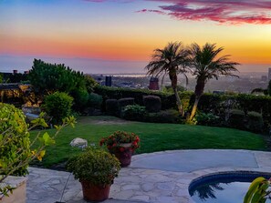 Un jardín ajardinado con piscina, plantas en macetas y vistas del horizonte de la ciudad al atardecer.