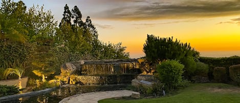 A garden with a stone pathway leading to a bridge over a water feature, surrounded by greenery and a sunset sky.