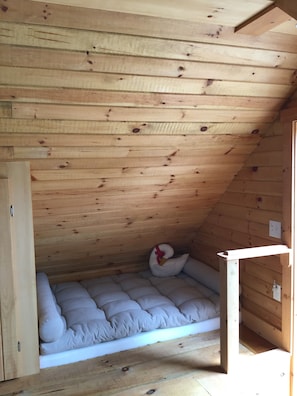 A small, cozy room with wooden slats on the ceiling, a white cushioned bench, and a red decorative pillow.