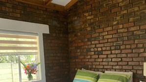 A bedroom with a brick wall, a bed with striped bedding, a bedside table with a lamp, and a window with a view of greenery.