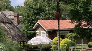 A swimming pool surrounded by a well-maintained garden with a patio table and chairs, a thatched-roof structure, and a brick house with a red roof in the background.