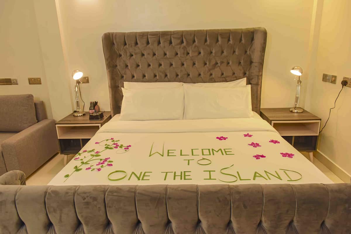 A neatly made bed with a "Welcome to One the Island" sign, two bedside lamps, and a grey upholstered headboard.