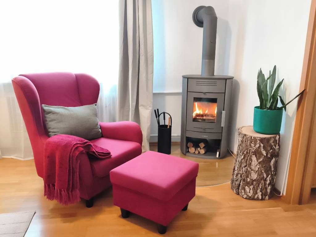 A cozy living room with a red armchair, a matching ottoman, a wood-burning stove, a potted plant, and a wooden stump.