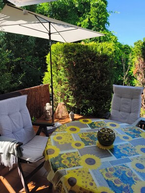 Un patio al aire libre con dos sillas blancas acolchadas, una mesa cubierta con mantel de estampado de girasoles y un gran paraguas.