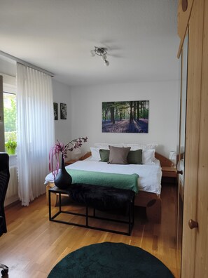A bedroom with a bed, a nightstand, a bedside table with a vase, a window with curtains, and a painting of a forest. - Sartorius Vacation Apartment, Vacation Apartment at Hemsberg (Bensheim)
