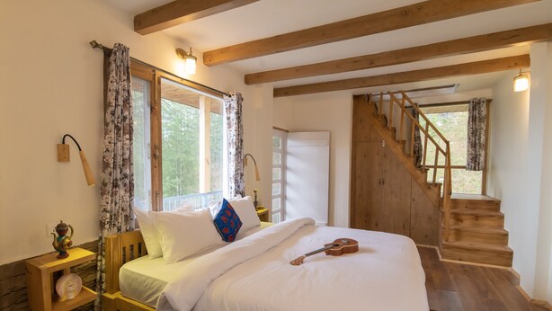 A bedroom with a wooden bed, white bedding, a guitar, a wooden nightstand with a lamp, and a staircase leading to another room. - Jomotel Dayaar (Theog)
