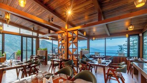 A spacious dining area with wooden tables and chairs, large windows offering mountain views, and hanging pendant lights. - Jomotel Dayaar (Theog)