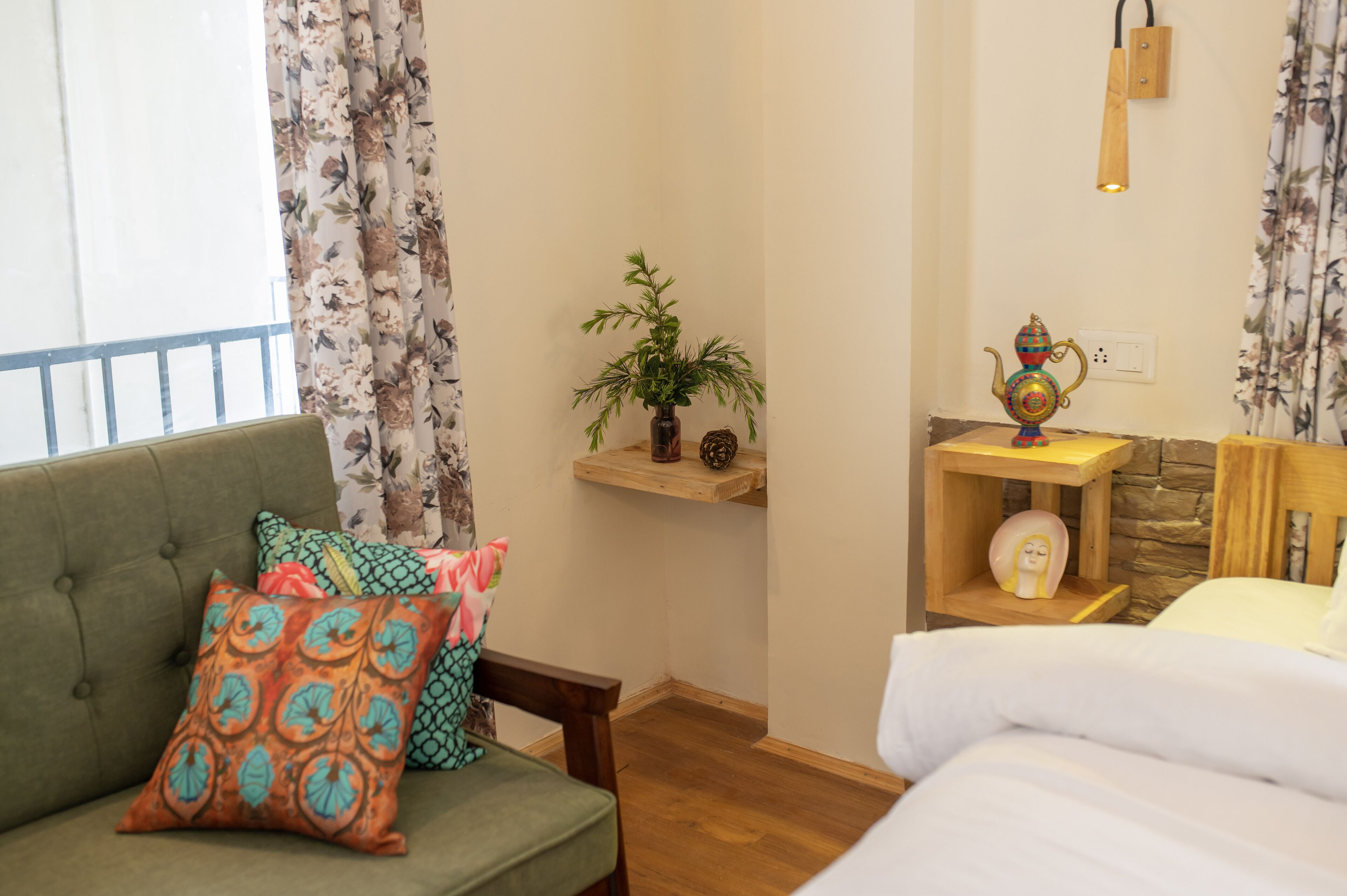 A cozy room with a green sofa, colorful pillows, a small plant on a shelf, a decorative lamp, and a bed with white bedding.