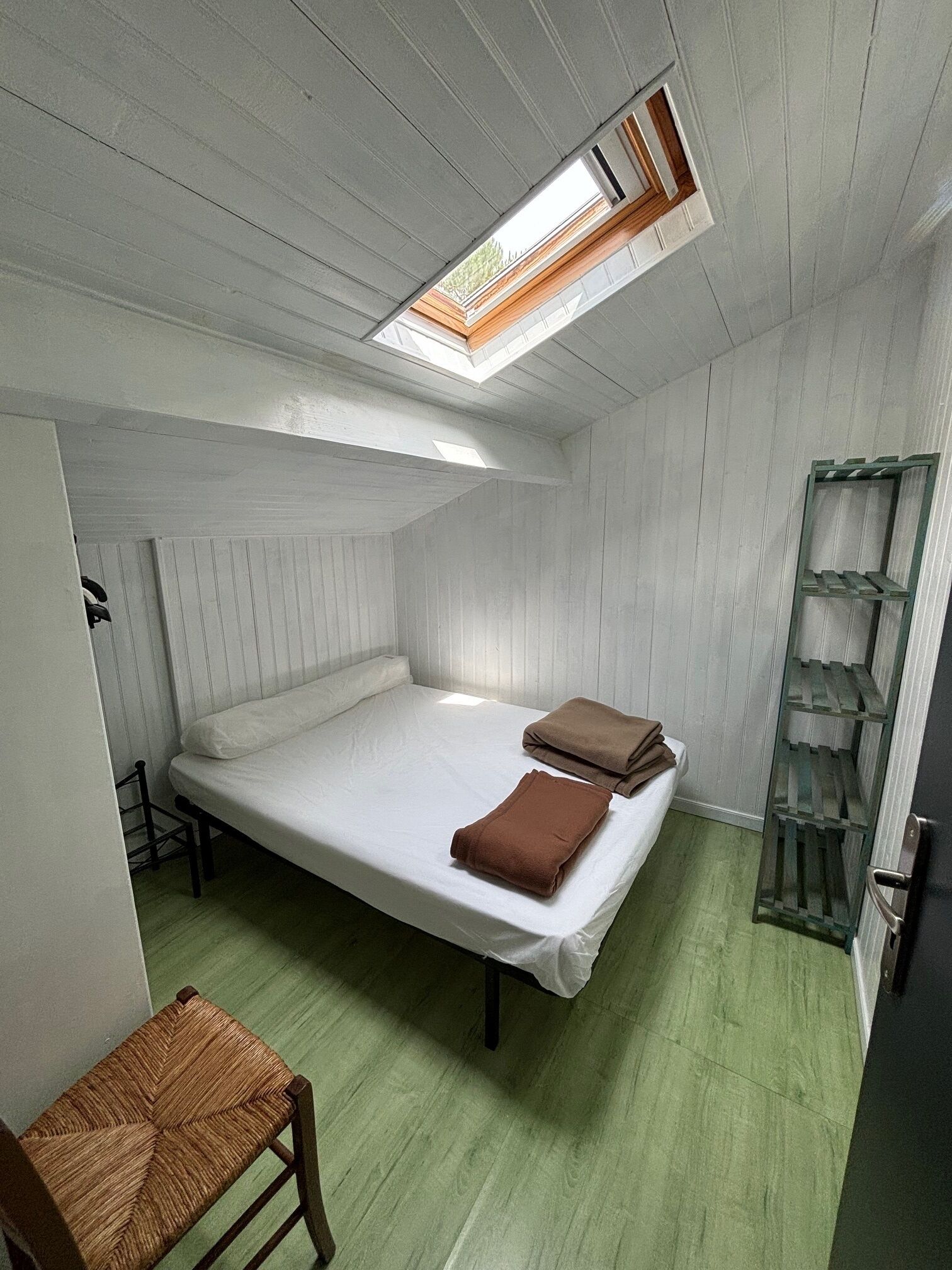 Ein kleines Zimmer mit einem Einzelbett, einem Oberlicht, einem Holzstuhl und einem Metallregal.