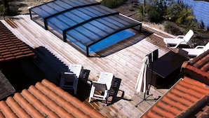 Un área de piscina con cerramiento de vidrio, terraza de madera y sillones de descanso.