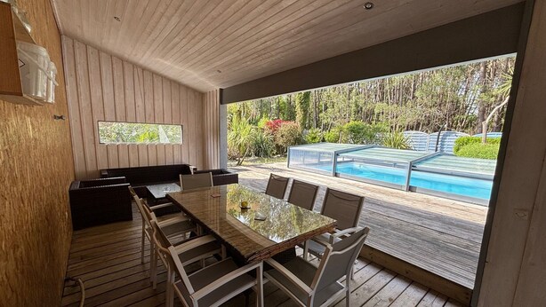 Un área de comedor al aire libre con una mesa y sillas de vidrio, piso de madera y vistas a una piscina y jardín.