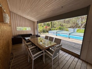 An outdoor dining area with a glass table and chairs, wooden flooring, and a view of a swimming pool and garden. - Near Soulac-sur-Mer, house for 6 pers private heated indoor pool (Grayan-et-l'Hôpital)