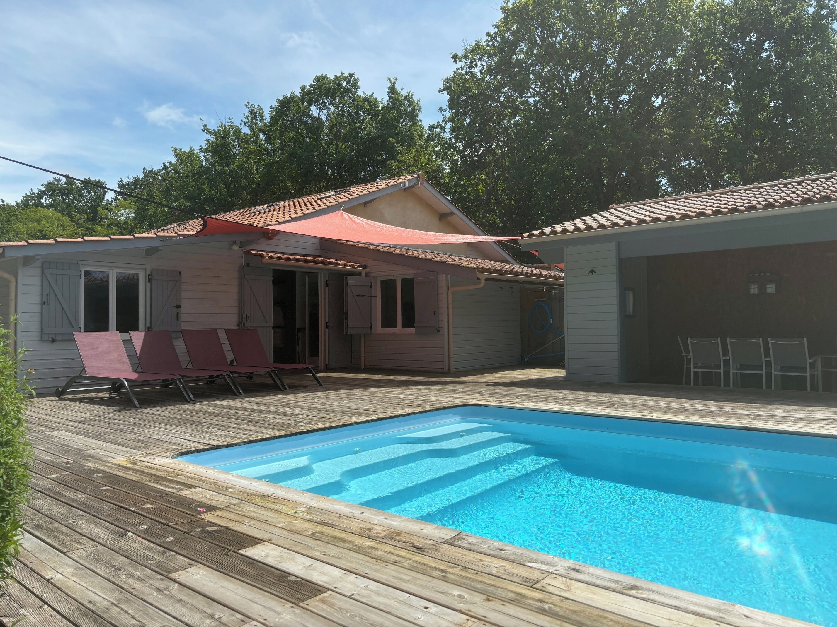 A pool area with a wooden deck, a covered seating area with chairs, and a building with a tiled roof.
