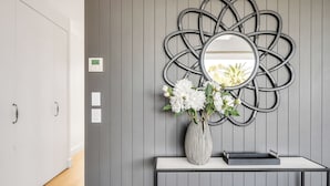 Una habitación moderna con pared gris, un espejo metálico decorativo, un jarrón blanco con flores y una bandeja negra sobre una consola blanca.