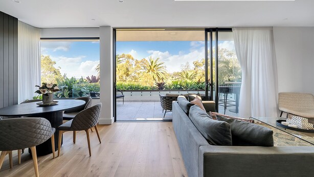 Un salón moderno con sofá, mesa de comedor y sillas, un balcón con vistas a zonas verdes y palmeras, y un amplio ventanal corredero.