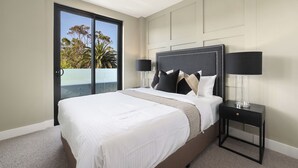 Habitación de hotel con una cama grande, una mesita de noche con lámpara y un ventanal corredera que da a un balcón con vistas a árboles y al cielo.