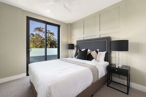A hotel room with a large bed, a nightstand with a lamp, and a sliding glass door leading to a balcony with a view of trees and the sky.