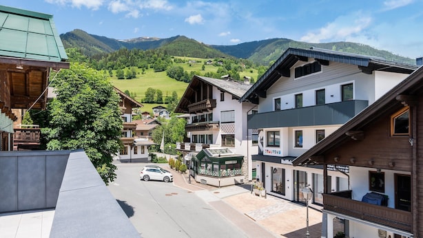 Straßenansicht einer Stadt mit traditionellen Holzbauten, einem auf der Straße geparkten Auto und einem Blick auf grüne Hügel im Hintergrund.