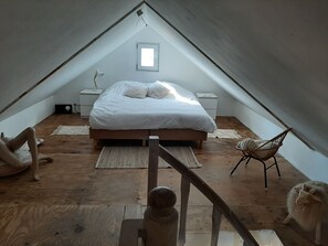 A cozy attic bedroom with a sloped ceiling, a bed with white bedding, a small window, a nightstand, a chair, and a wooden floor.